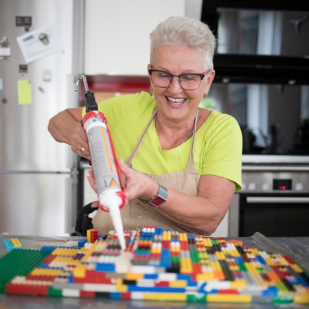 Eine 64-järhige Frau, mit grauen Haaren, Brille und einen Lächeln sitzt an einem Tisch und baut eine Rampe aus Legosteinen.