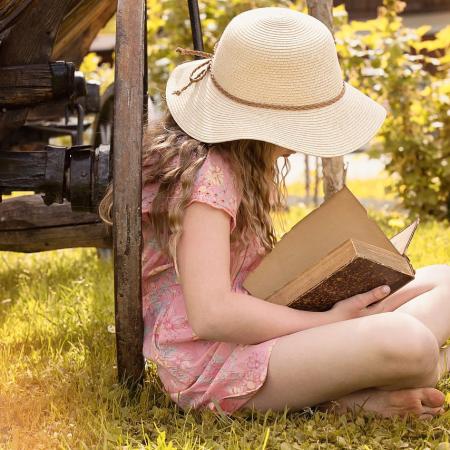 Ein Mädchen mit Hut sitz an einen Holzwagen gelehnt im Schneidersitz auf der Wiese und liest in einem Buch