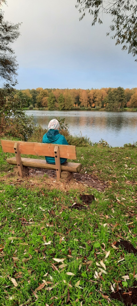 Frau mit grauer Mütze und blauer Jacke, die von hinten sitzend auf einer Bank fotografiert wurde. Sie sitzt an einem See