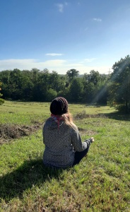 Eine Frau mit Mütze, die mit dem Rücken zur Kamera sitzt und meditiert. Sie sitzt auf einer Weise bei blauem Himmel und Sinnenschein